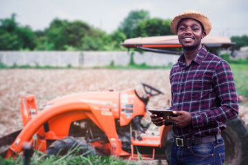 African farmer is using a tablet on the background of working tractor with a cultivator in the...
