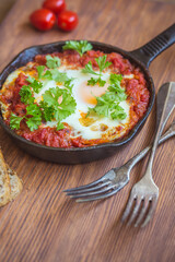 Shakshuka - fried eggs with vegetables in iron cast on wooden table