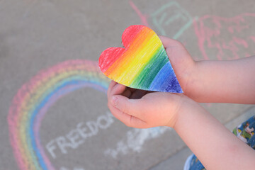 child holds in palms a paper heart painted in rainbow colors of lgbt community rainbow, chalk on pavement, month pride