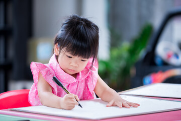 Cute child doing artwork on red desk. Asian girl drawing cartoon on paper. Happy kid learning at home. Children aged 4 years old.