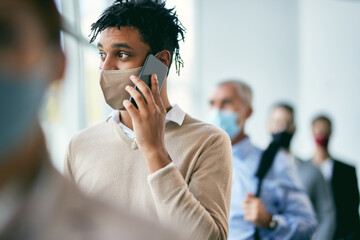 Young African American businessman making phone call while waiting in line and wearing face mask...