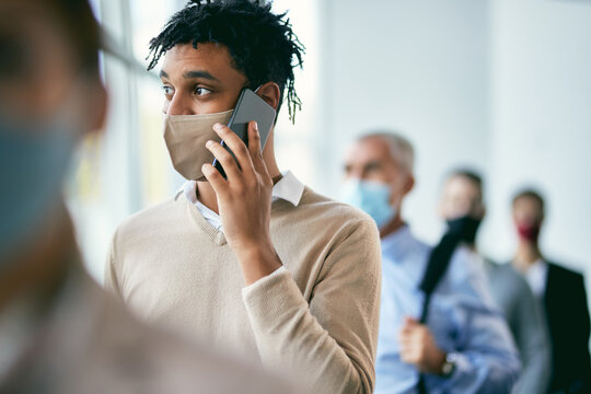Young African American Businessman Making Phone Call While Waiting In Line And Wearing Face Mask Due To Coronavirus Pandemic.