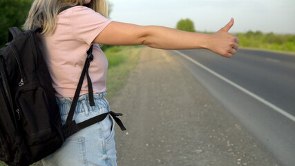 Fototapeta na wymiar A girl with a backpack stands by the road and catches the car with her thumb up. Hitchhiking. Stop the car with your hand