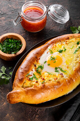  A traditional dish of Georgian cuisine, Adjarian khachapuri with cheese and egg and spicy tomato sauce adjika on the dark background close up vertical photo