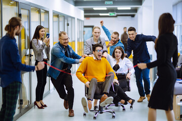 Young colleagues group having fun together, riding on chairs in office, diverse excited office...