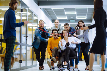 Young colleagues group having fun together, riding on chairs in office, diverse excited office...