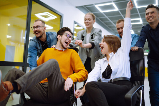 Young colleagues group having fun together, riding on chairs in office, diverse excited office workers enjoying break, laughing, engaged funny activity, celebrating corporate success.