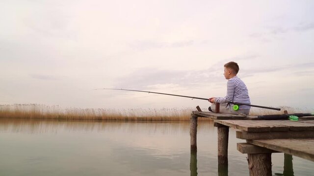A boy sits on a wooden bridge and catches a fish on fishing rod. 