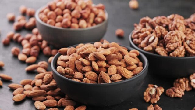 Assortment shelled grains of nuts, almonds, walnuts and hazelnuts rotate around its axis. Different nuts in the bowls on dark wooden background, high-calorie vegan food.