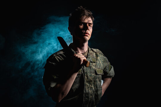 Portrait of a soldier with scars and combat coloring, holding a machete in the urns on a black background witch blue smoke