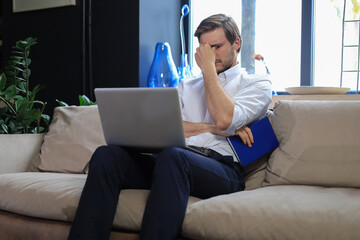 Fototapeta na wymiar Unhappy frustrated young male holding head by hand sitting with laptop on sofa.