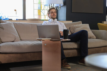 Tired young male sitting with closed eyes on sofa with laptop standing near him.