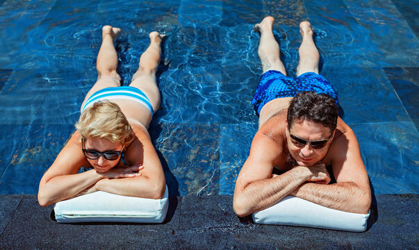 Successful Retirement Recreation, Summer Vacation Concept. Retired Mature Couple Enjoying Beautiful Sunny Day In Swimming Pool At Beach Club. Happy Senior Woman And Man Lying In Water At Poolside.