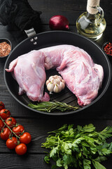 Raw rabbit meat with fresh vegetables, herbs and spices, in cast iron frying pan, on black wooden table background
