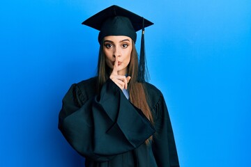 Beautiful brunette young woman wearing graduation cap and ceremony robe asking to be quiet with finger on lips. silence and secret concept.