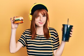 Redhead young woman eating a burger and drinking soda wearing headphones winking looking at the...