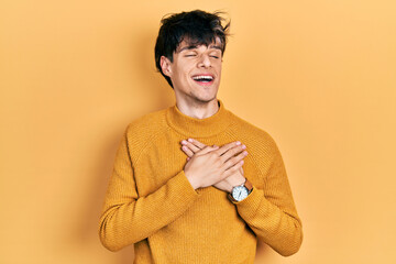 Handsome hipster young man wearing casual yellow sweater smiling with hands on chest with closed...