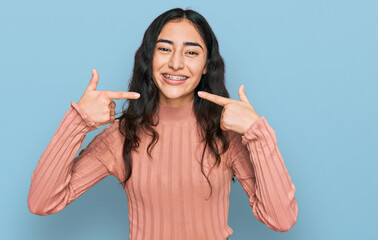 Hispanic teenager girl with dental braces wearing casual clothes smiling cheerful showing and pointing with fingers teeth and mouth. dental health concept. - obrazy, fototapety, plakaty
