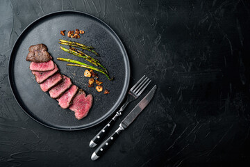 Juicy steak medium rare beef Filet Mignon or Eye Fillet, with onion and asparagus, on plate, with meat knife and fork, on black stone background, top view flat lay, with copy space for text - obrazy, fototapety, plakaty