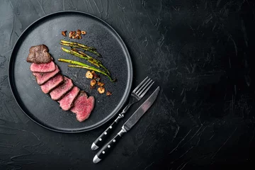 Poster Juicy steak medium rare beef Filet Mignon or Eye Fillet, with onion and asparagus, on plate, with meat knife and fork, on black stone background, top view flat lay, with copy space for text © Ilia Nesolenyi