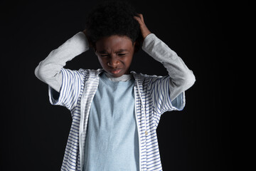 Portrait of little African boy holding his head with two hands with a face of pain or stress or headache or worry and confusion with hoodies sweatshirt standing on black background. Copy space