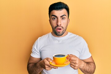 Young hispanic man drinking a cup of coffee in shock face, looking skeptical and sarcastic, surprised with open mouth