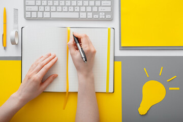 Female hands writing an idea goal into blank notebook on desktop in workspace. Woman writes list of ideas in notebook on gray yellow background. Yellow light bulb idea metaphor.