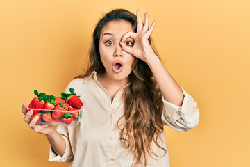 Young hispanic girl holding strawberries doing ok gesture shocked with surprised face, eye looking through fingers. unbelieving expression.