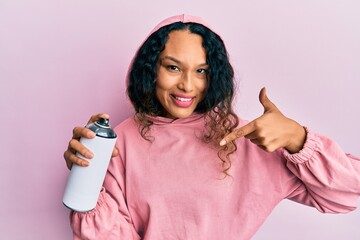 Young latin woman wearing sweatshirt holding graffiti spray pointing finger to one self smiling...