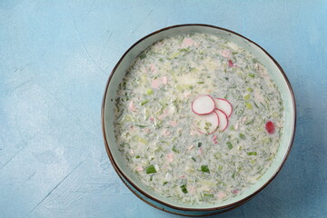 Cold soup okroshka with sausage, potato, egg, radish, cucumber, greens and kvass in a bowl, bread on wicker napkin. Traditional Russian summer cold soup with kvass