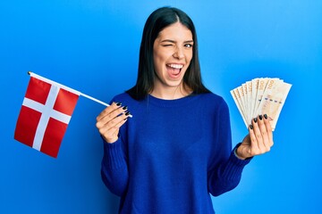 Young hispanic woman holding denmark flag and krone banknotes winking looking at the camera with...