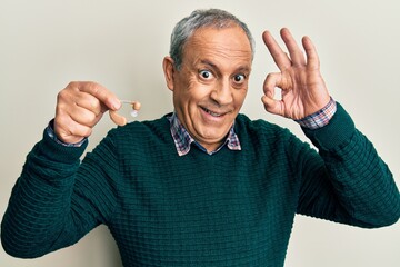 Handsome senior man with grey hair holding medical hearing aid doing ok sign with fingers, smiling...