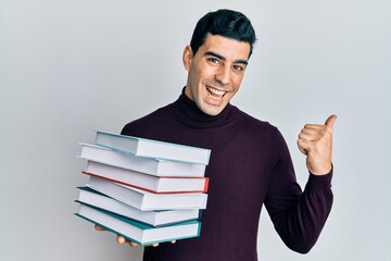 Handsome hispanic man holding a pile of books pointing thumb up to the side smiling happy with open mouth
