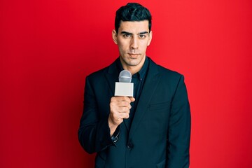 Handsome hispanic man holding reporter microphone thinking attitude and sober expression looking...