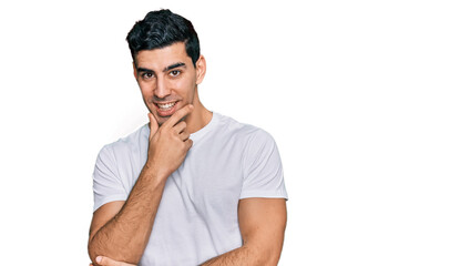 Handsome hispanic man wearing casual white t shirt looking confident at the camera with smile with crossed arms and hand raised on chin. thinking positive.