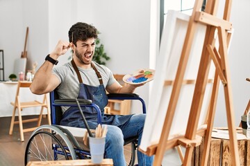 Young hispanic man sitting on wheelchair painting at art studio angry and mad raising fist frustrated and furious while shouting with anger. rage and aggressive concept.