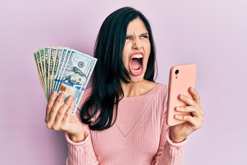 Young hispanic woman using smartphone holding united states dollar banknotes angry and mad screaming frustrated and furious, shouting with anger. rage and aggressive concept.