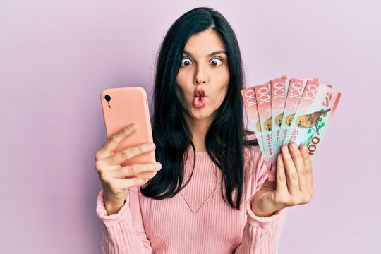 Young hispanic woman using smartphone holding new zealand dollars banknotes making fish face with mouth and squinting eyes, crazy and comical.