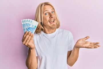 Caucasian young man with long hair holding 50 polish zloty banknotes celebrating achievement with happy smile and winner expression with raised hand