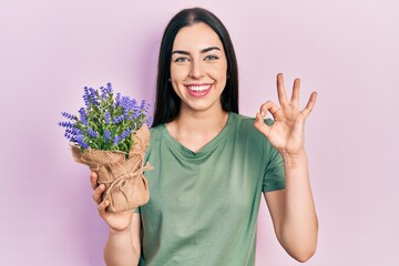 Beautiful woman with blue eyes holding lavender plant doing ok sign with fingers, smiling friendly gesturing excellent symbol