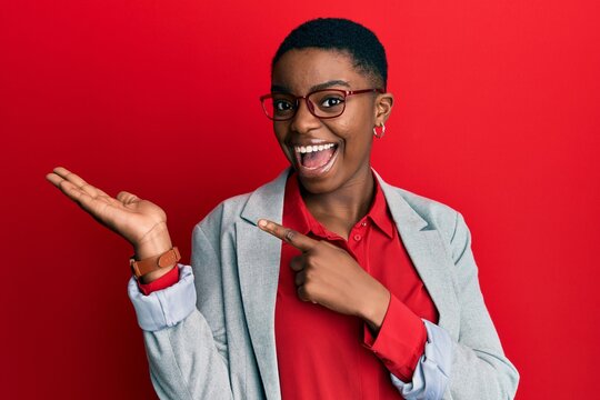 Young african american woman wearing business jacket and glasses amazed and smiling to the camera while presenting with hand and pointing with finger.
