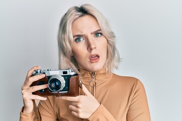 Young blonde girl holding vintage camera in shock face, looking skeptical and sarcastic, surprised with open mouth