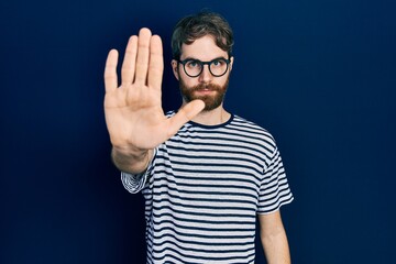 Caucasian man with beard wearing striped t shirt and glasses doing stop sing with palm of the hand. warning expression with negative and serious gesture on the face.