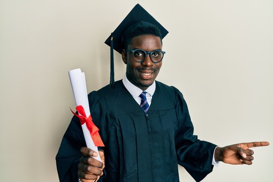 Handsome black man wearing graduation cap and ceremony robe holding diploma smiling happy pointing with hand and finger to the side