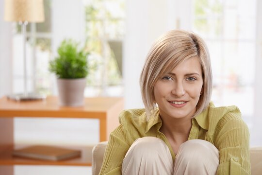 Portrait of smiling woman sitting with arms around knees in living room, looking at camera.