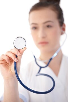 Young woman in lab coat holding stethoscope towards camera, looking serious. Isolated on white.