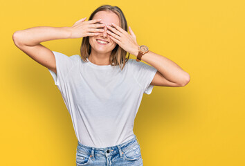 Beautiful young blonde woman wearing casual white t shirt covering eyes with hands smiling cheerful and funny. blind concept.
