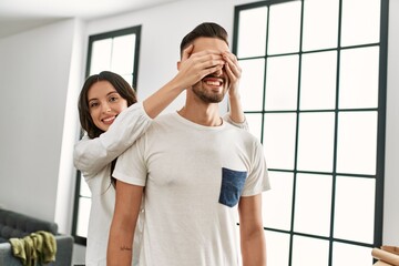 Young hispanic woman surprising man putting hands on eyes at new home.