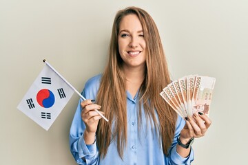 Young blonde woman holding south korea flag and won banknotes winking looking at the camera with...