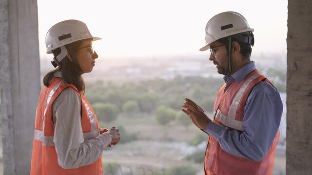 close view arc shot of young indian asian male and female civil engineers having a conversation together standing on top of under development building near construction site